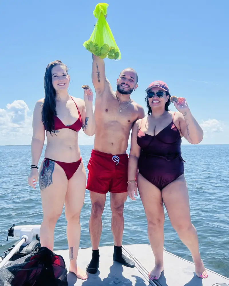Three adults on the bow of a boat holding up a mesh bag of bay scallops