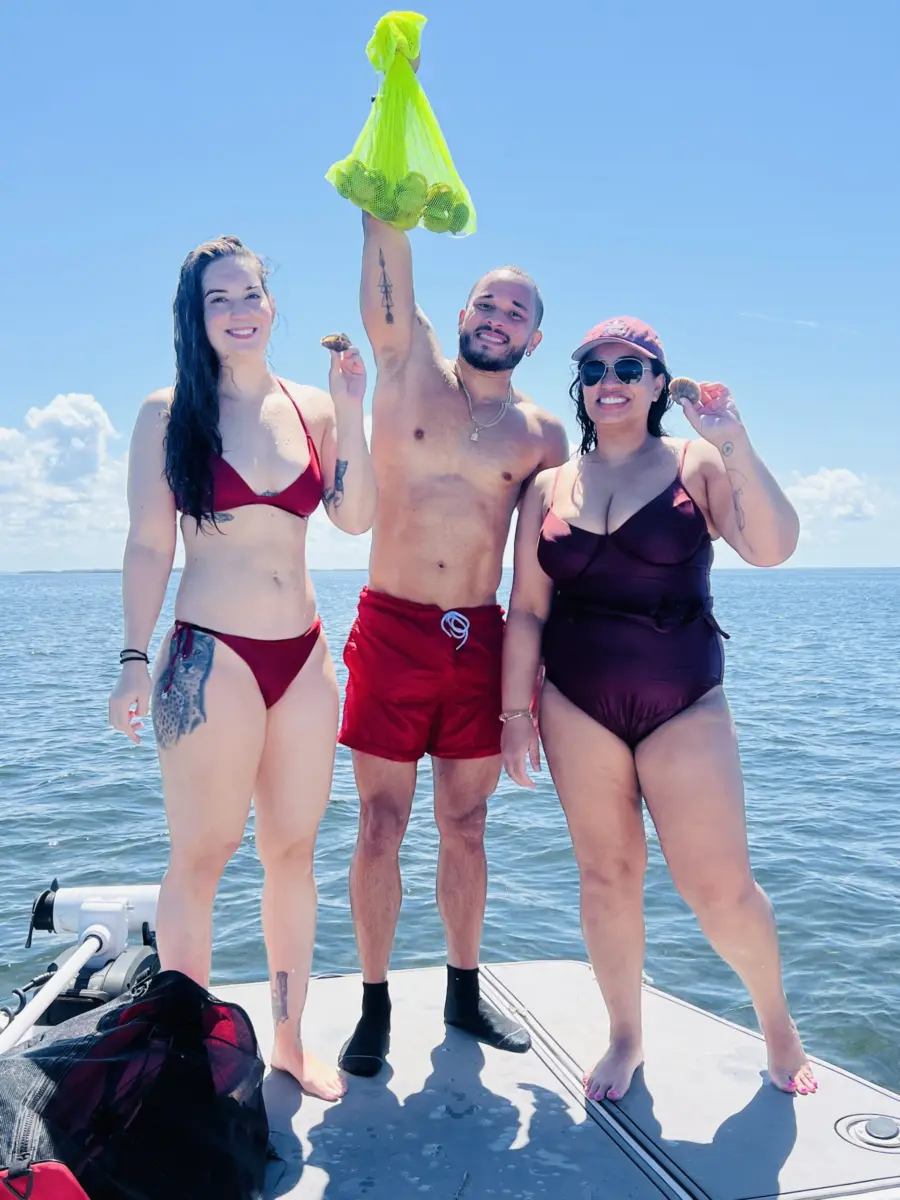 Three adults on the bow of a boat holding up a mesh bag of bay scallops