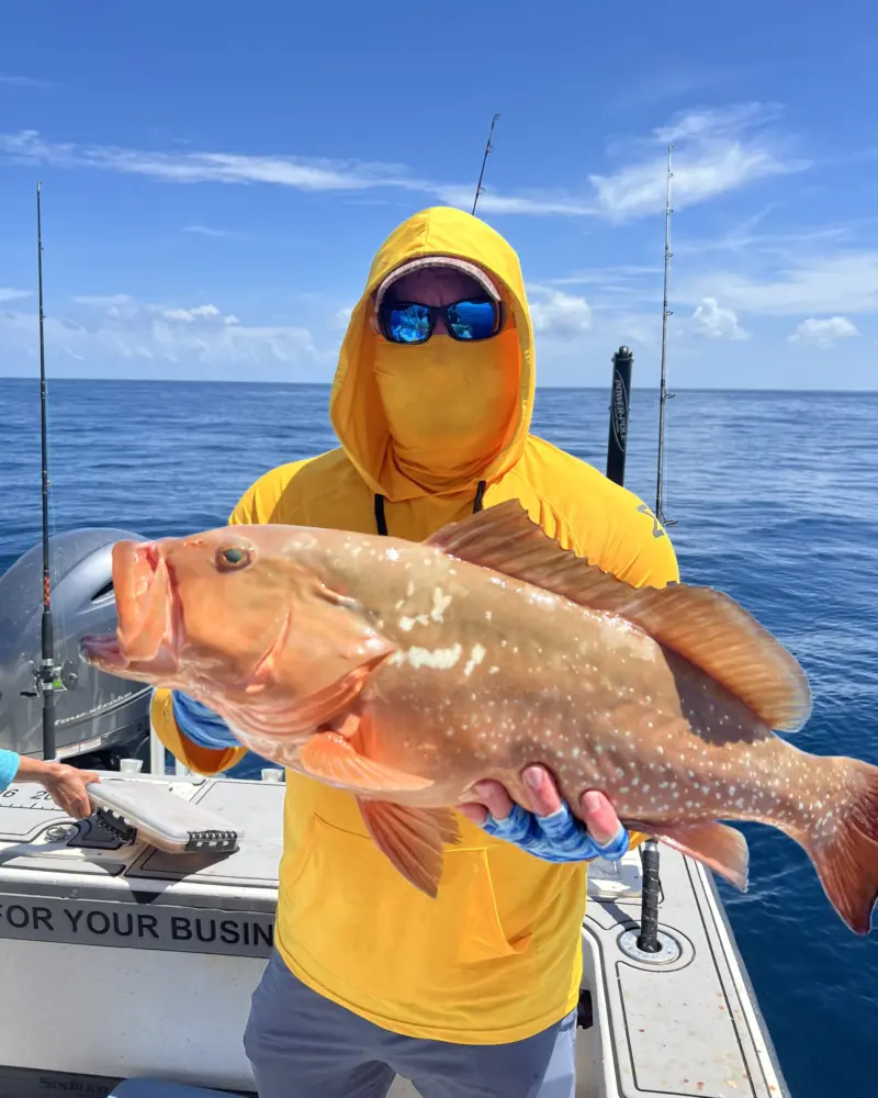 Angler in a yellow hoodie holding a red grouper broadside with blue Gulf water behind