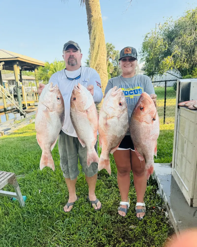 Man and teenage girl each holding two large red snapper on the lawn beside the Native Charters dock