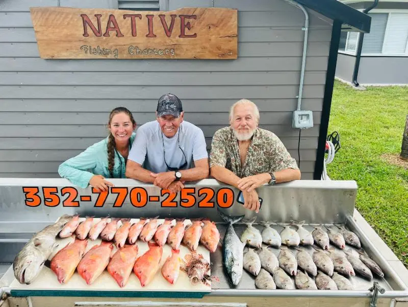 A woman and two men lean on the Native Fishing Charters cleaning table in front of a spread of snapper, hogfish and grunts