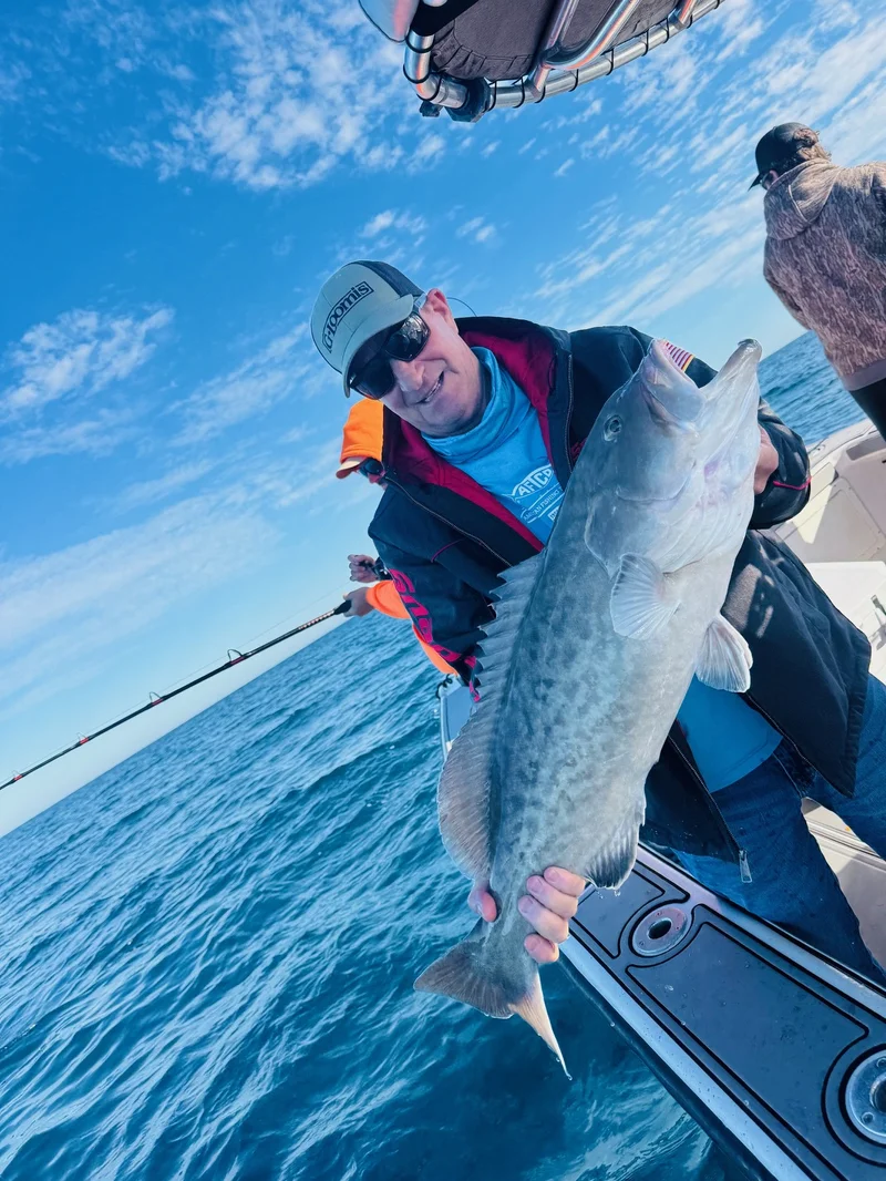 An angler in sunglasses holds a large grouper aboard a boat with blue sky and open water behind him