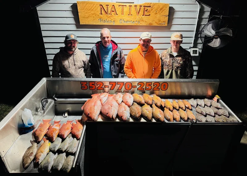 Four anglers stand behind a fish cleaning table stacked with red snapper and hogfish under a wooden Native Fishing Charters sign at night