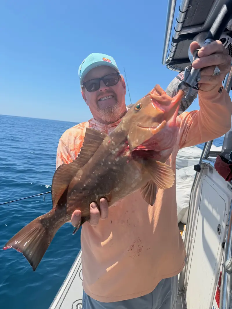 Man in a peach shirt holding a red grouper on a fish grip under a boat hardtop